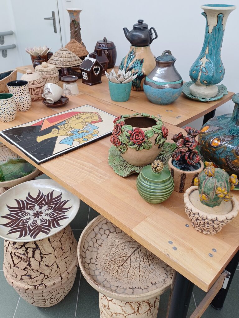 Zdjęcie przedstawia stół z różnorodnymi wyrobami ceramicznymi i rękodziełem. Wśród nich znajdują się wazony o różnych kształtach i zdobieniach (jeden turkusowy z falami, inny brązowy z reliefami), doniczki z roślinami (sukulentami i kaktusem), ozdobne naczynia, dzbanki, puszki oraz tace i podstawki. Na pierwszym planie po lewej stronie widać okrągłą tacę z ciemnobrązowym wzorem przypominającym śnieżynkę lub kwiat. Na środku stołu leży obraz z postaciami w ludowym stylu. W tle widoczne są inne ceramiczne przedmioty. Wszystkie wyroby charakteryzują się unikalnym wzornictwem i kolorystyką.