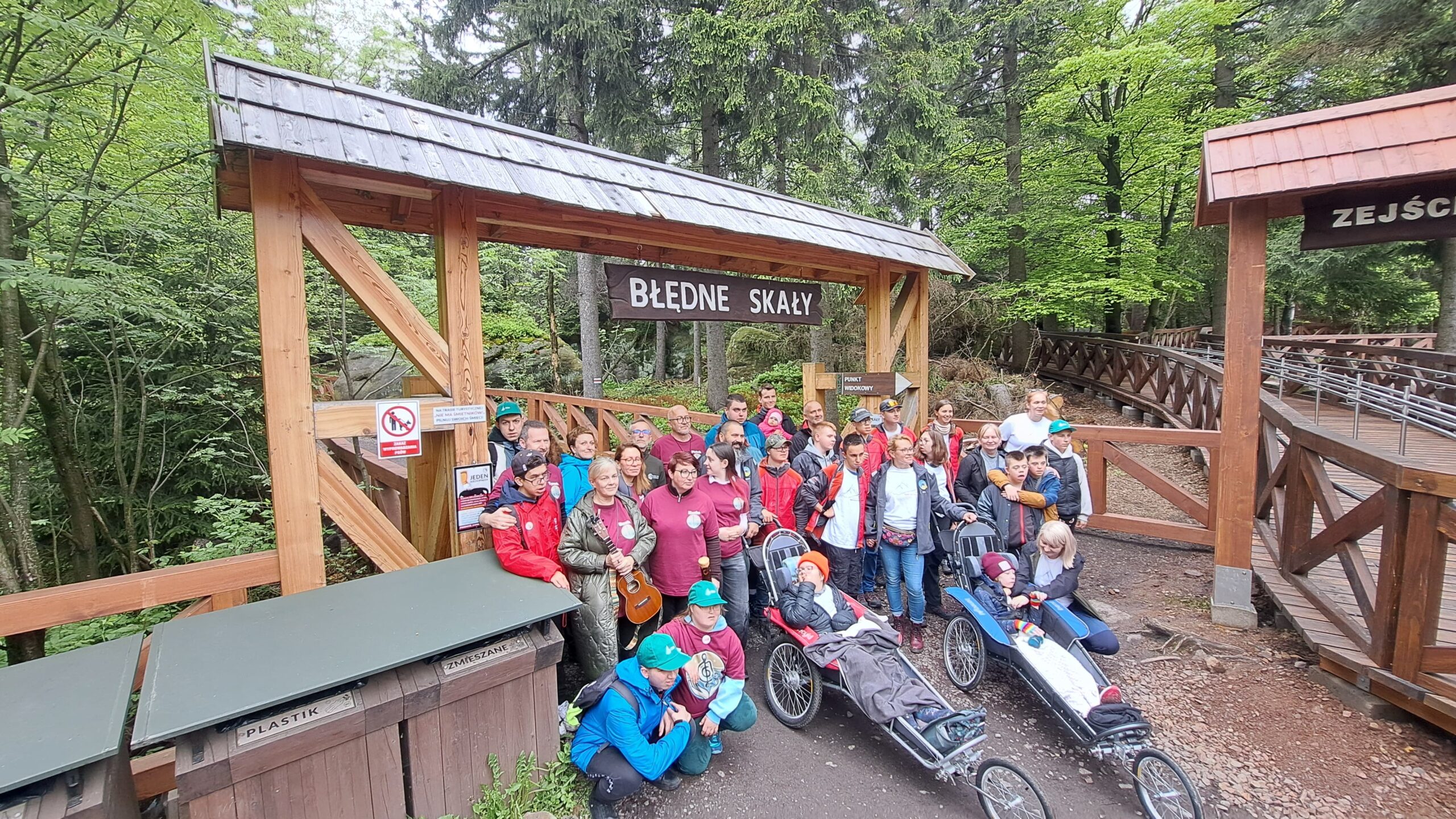 Duża grupa ludzi, w tym dorośli i młodzież, pozuje do zdjęcia na zalesionym obszarze, prawdopodobnie przy wejściu na szlak turystyczny. W centrum kadru znajduje się drewniana konstrukcja z dachem i tabliczką z napisem „BŁĘDNE SKAŁY”. Po lewej stronie widać pojemniki do segregacji śmieci, w tym jeden opisany jako „PLASTIK”. Po prawej stronie zdjęcia widoczna jest drewniana balustrada i fragment innej drewnianej konstrukcji. Część osób siedzi, a dwie z nich znajdują się w specjalnych wózkach terenowych. W tle rosną wysokie drzewa z zielonymi liśćmi.