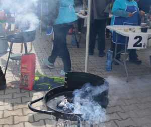 Na brukowanej powierzchni stoi kilka rozpalonych, czarnych grillów na węgiel drzewny, z których unosi się gęsty, szary dym. W pierwszym planie widoczny jest okrągły grill z widocznymi żarzącymi się brykietami węgla. Obok niego leży czerwona torba z napisem "Brykiet drzewny". W tle widać fragmenty białych stolików z numerami, butelki wody mineralnej oraz rozmyte postacie ludzi. Dym tworzy zamgloną atmosferę, częściowo zasłaniając otoczenie.