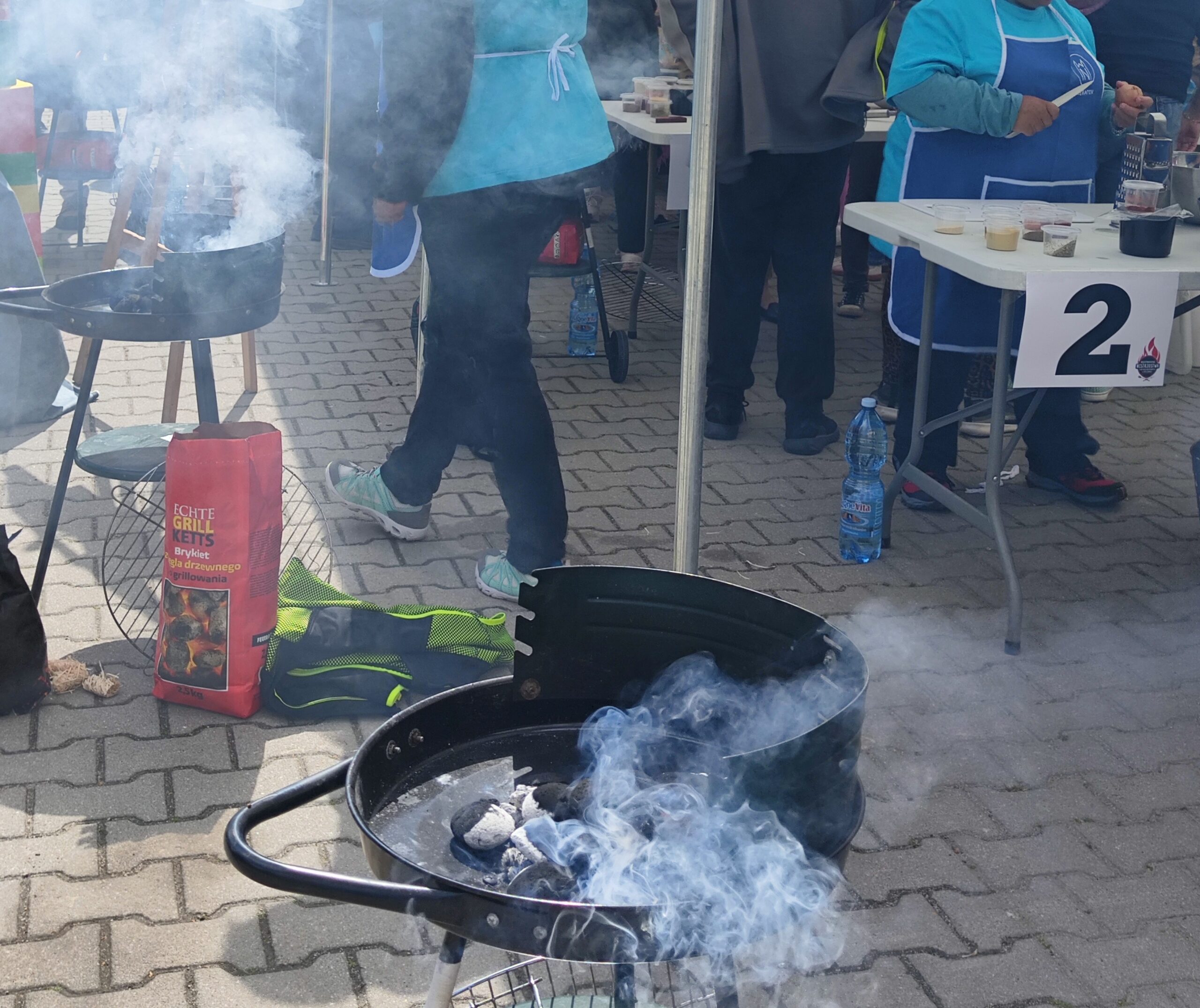 Na brukowanej powierzchni stoi kilka rozpalonych, czarnych grillów na węgiel drzewny, z których unosi się gęsty, szary dym. W pierwszym planie widoczny jest okrągły grill z widocznymi żarzącymi się brykietami węgla. Obok niego leży czerwona torba z napisem "Brykiet drzewny". W tle widać fragmenty białych stolików z numerami, butelki wody mineralnej oraz rozmyte postacie ludzi. Dym tworzy zamgloną atmosferę, częściowo zasłaniając otoczenie.