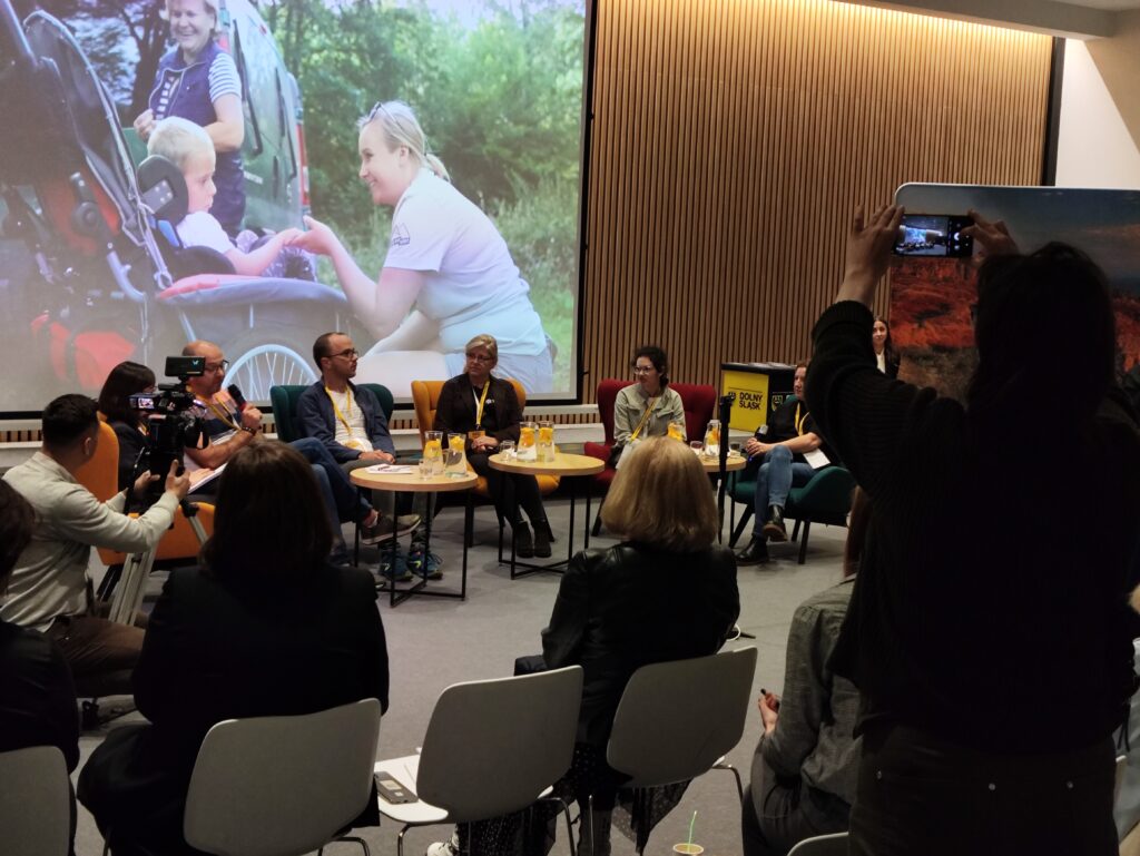 Wnętrze sali z rzędami krzeseł zwróconych w stronę ekranu projekcyjnego, na którym wyświetlane jest niewyraźne zdjęcie. Kilka osób siedzi przy stolikach, a jedna osoba stoi z podniesionym telefonem, prawdopodobnie robiąc zdjęcie ekranu. Ściana za ekranem jest pokryta drewnianymi panelami.