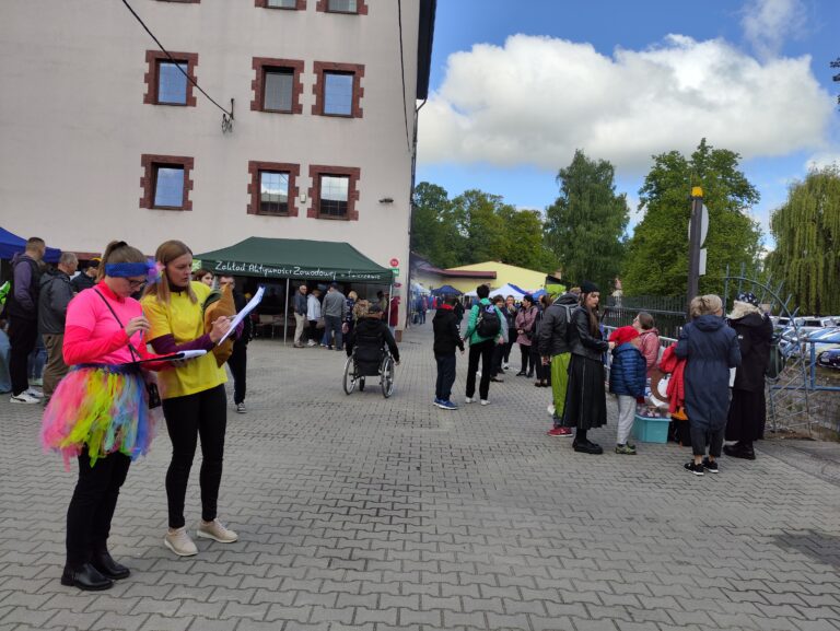 Dwie młode kobiety stoją na brukowanej powierzchni i piszą na clipboardach. Wokół nich zgromadzony jest tłum ludzi, a w tle widać budynek i festynowe namioty.