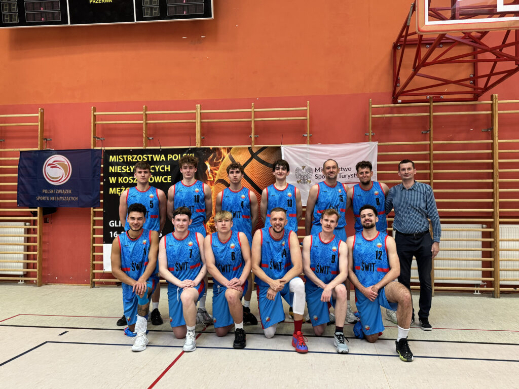 Drużyna koszykówki mężczyzn, złożona z dziesięciu zawodników i jednego trenera, pozuje do zdjęcia. Wszyscy ubrani są w niebieskie koszulki z napisem "ŚWIT" i niebieskie spodenki. Za nimi widać transparenty z napisami "MISTRZOSTWA POLSKI NIESŁYSZĄCYCH W KOSZYKÓWCE MĘŻCZYZN" oraz "GLIWICE 16-18.05.2025".