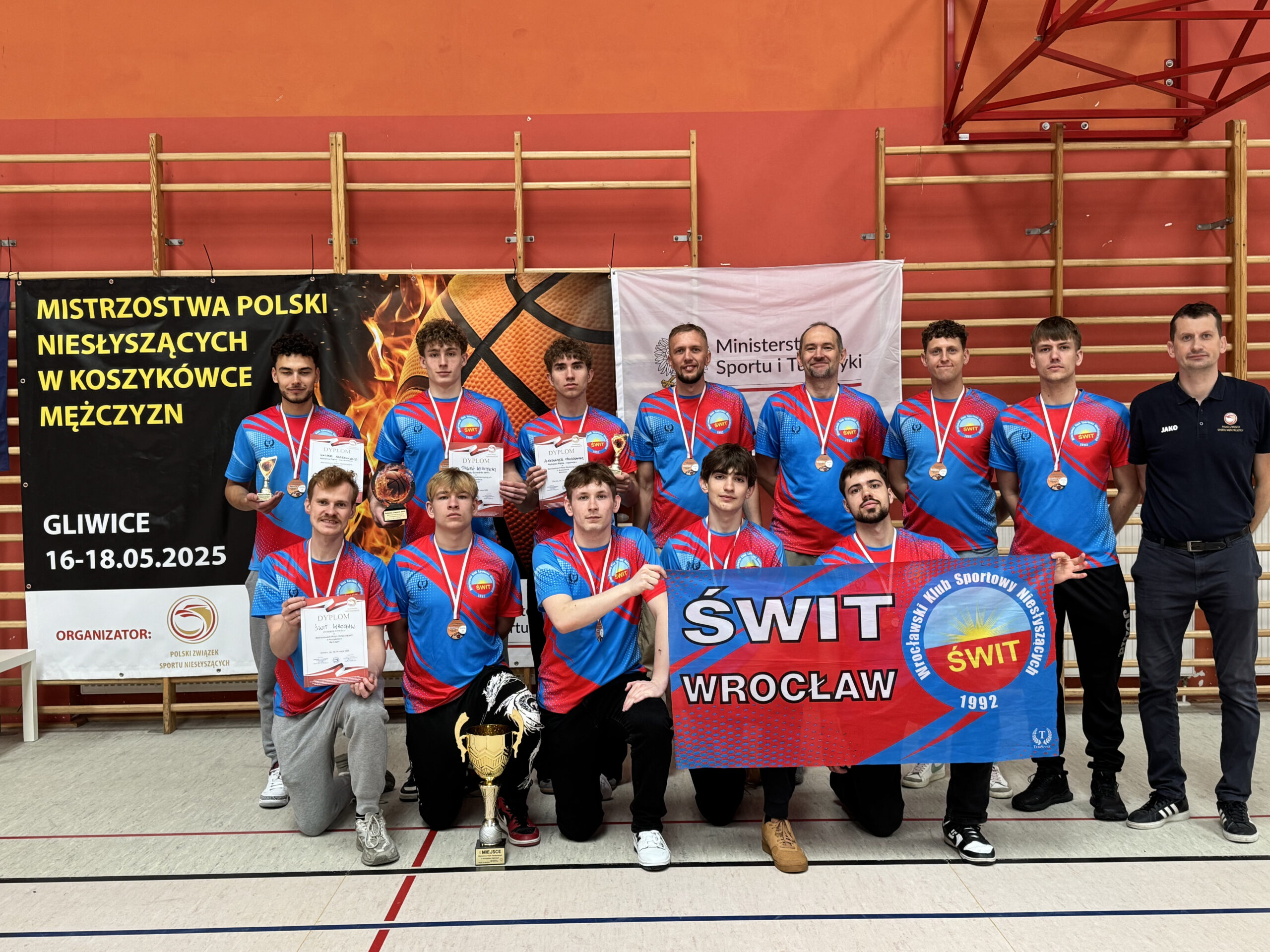 Drużyna koszykówki mężczyzn składa się z jedenastu zawodników i jednego trenera. Wszyscy ubrani są w niebiesko-czerwone stroje sportowe i mają na szyjach medale. Pośrodku stoi duży złoty puchar. Za nimi widać transparenty z napisami "MISTRZOSTWA POLSKI NIESŁYSZĄCYCH W KOSZYKÓWCE MĘŻCZYZN", "GLIWICE 16-18.05.2025" oraz "ŚWIT WROCŁAW".