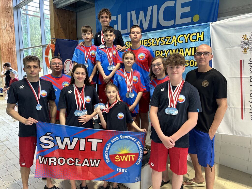 Grupa młodych pływaków i dorosłych (prawdopodobnie trenerzy) pozuje na tle basenu. Większość osób ma na szyjach medale. Na pierwszym planie widać transparent z napisem "ŚWIT WROCŁAW" oraz logiem Wrocławskiego Klubu Sportowego Niesłyszących ŚWIT. W tle widać banner z napisem "GLIWICE" i "MISTRZOSTWA POLSKI NIESŁYSZĄCYCH W PŁYWANIU".