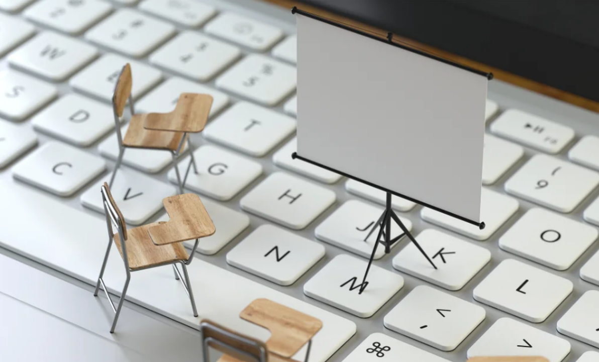 Miniaturowe biurka studenckie i ekran projekcyjny ustawione na klawiaturze laptopa, symbolizujące szkolenia online.