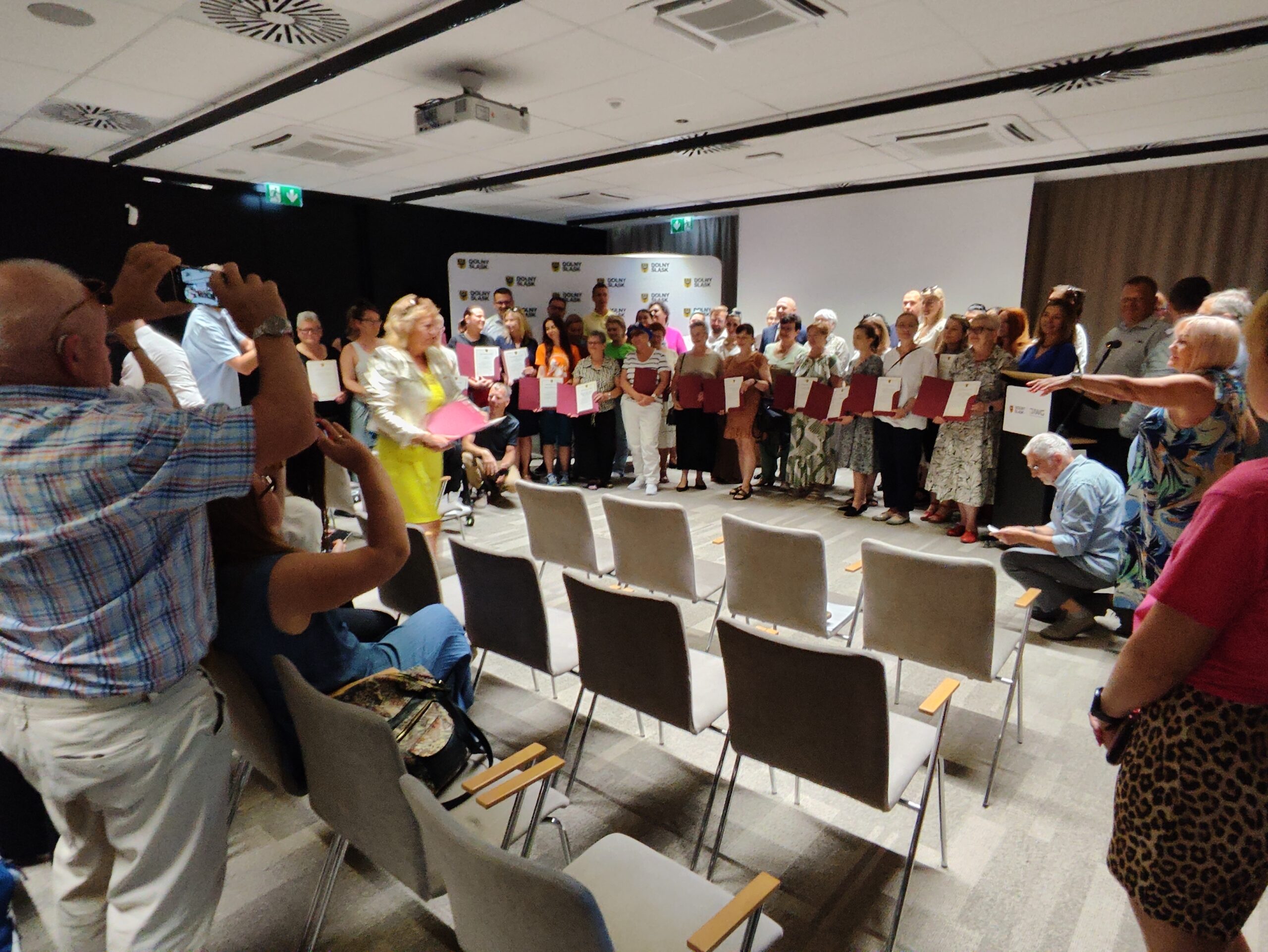 Obraz przedstawia dużą grupę dorosłych osób zgromadzonych w pomieszczeniu. Większość z nich stoi na końcu sali, niektóre trzymając certyfikaty lub dokumenty. Wyglądają, jakby brali udział w ceremonii lub wydarzeniu. Na pierwszym planie widać kilka pustych krzeseł i osoby siedzące lub stojące, niektóre robią zdjęcia lub nagrywają wideo. Pomieszczenie ma białe ściany, ciemną podłogę i oświetlenie sufitowe, a także duży ekran na ścianie w tle.