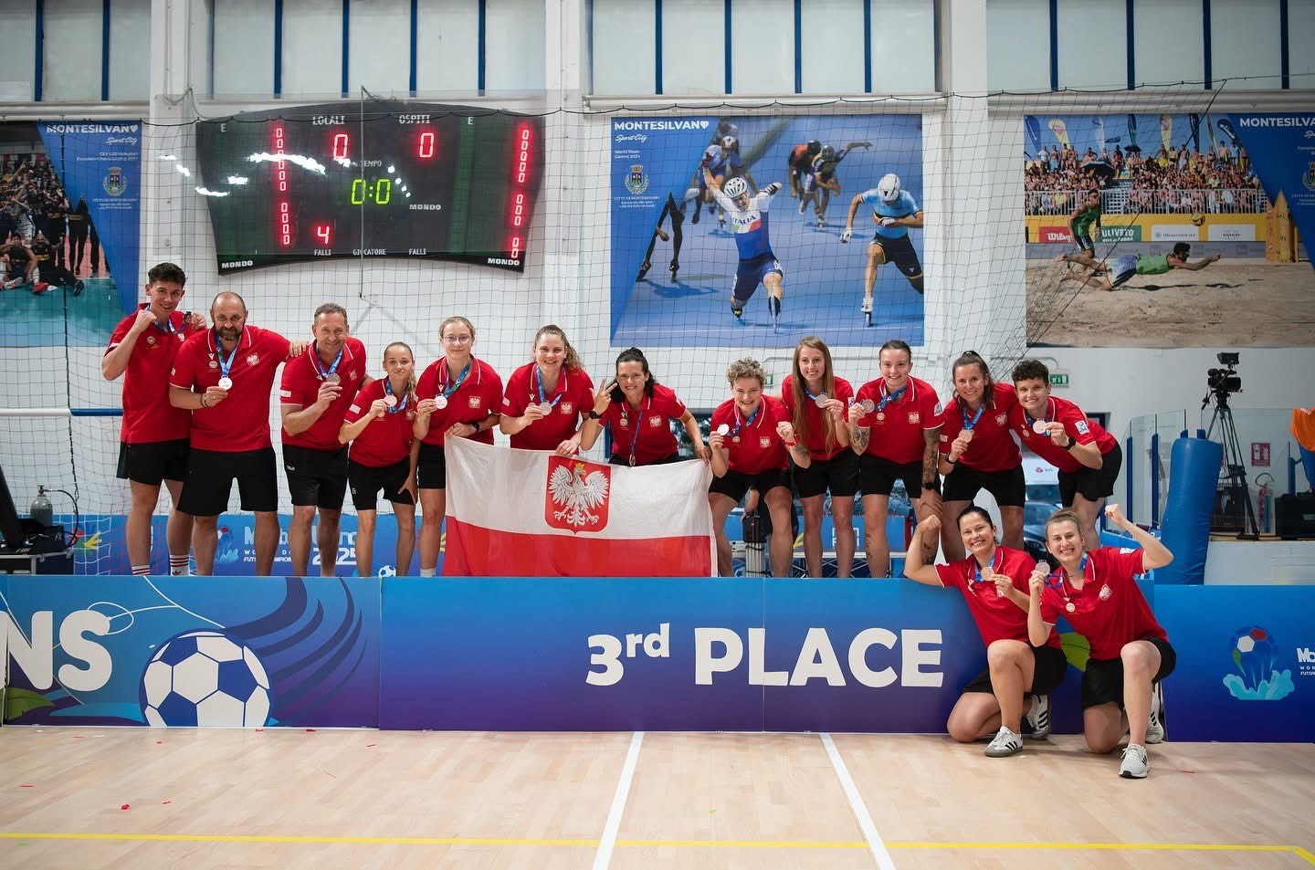 Zdjęcie przedstawia grupę młodych sportowców w czerwonych koszulkach, z których większość ma medale na szyjach, stojących lub klęczących na boisku sportowym. Trzymają polską flagę. Za nimi widać tablicę wyników i dużą grafikę przedstawiającą sportowców w akcji. Na dole zdjęcia widnieje napis "3rd PLACE".