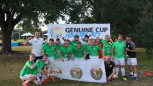 Grupa zawodników drużyny piłkarskiej WKS Śląsk Wrocław 'Bez Barier Futsal' pozuje radośnie przed banerem turnieju 'Genuine Cup Houston 2025'. Wszyscy ubrani w zielone koszulki i białe spodenki, niektórzy unoszą pięści w geście zwycięstwa. Na pierwszym planie trzymana jest flaga drużyny z herbem WKS Śląsk i napisem 'Bez Barier Futsal'. W tle widać drzewa, namioty oraz boiska sportowe. Atmosfera na zdjęciu jest pełna radości i dumy.