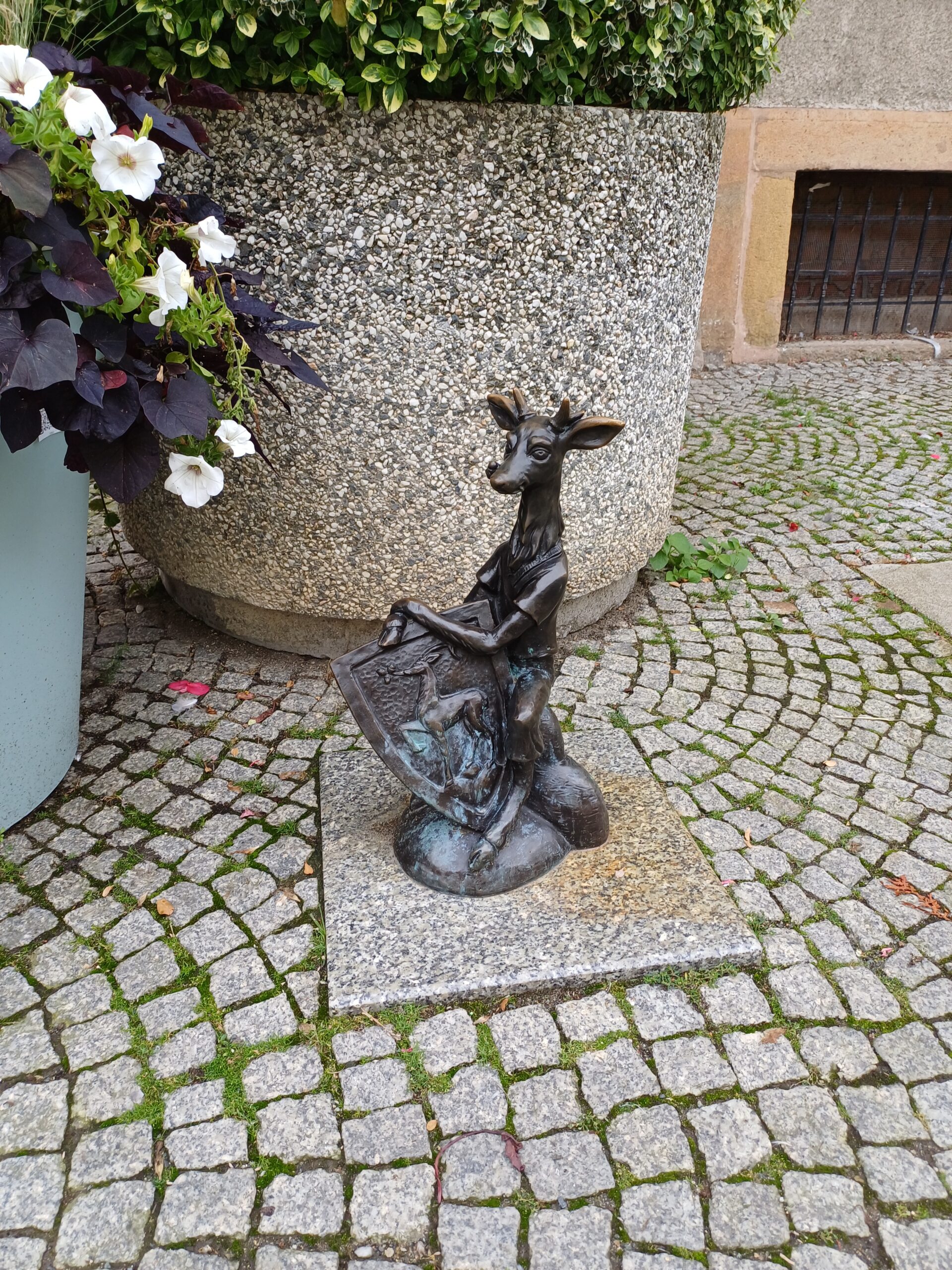 Miedziana rzeźba, przedstawiająca jelonka, siedzącego i trzymającego tarczę z herbem. W tle widoczna jest brukowana kostka, duża donica z roślinami oraz fragment okratowanego okna.