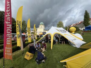 Widok na plenerową imprezę odbywającą się na trawniku pod zachmurzonym niebem. Na pierwszym planie widać dwie osoby siedzące na żółtych sześcianach i dwie siedzące na trawie. W tle stoją namioty, flagi i stoiska, a nad wszystkim góruje okazały budynek z kopułą Uzdrowiska Lądek Zdrój oraz drzewa. Na flagach, sześcianach i namiotach widnieje napis "DOLNY ŚLĄSK".