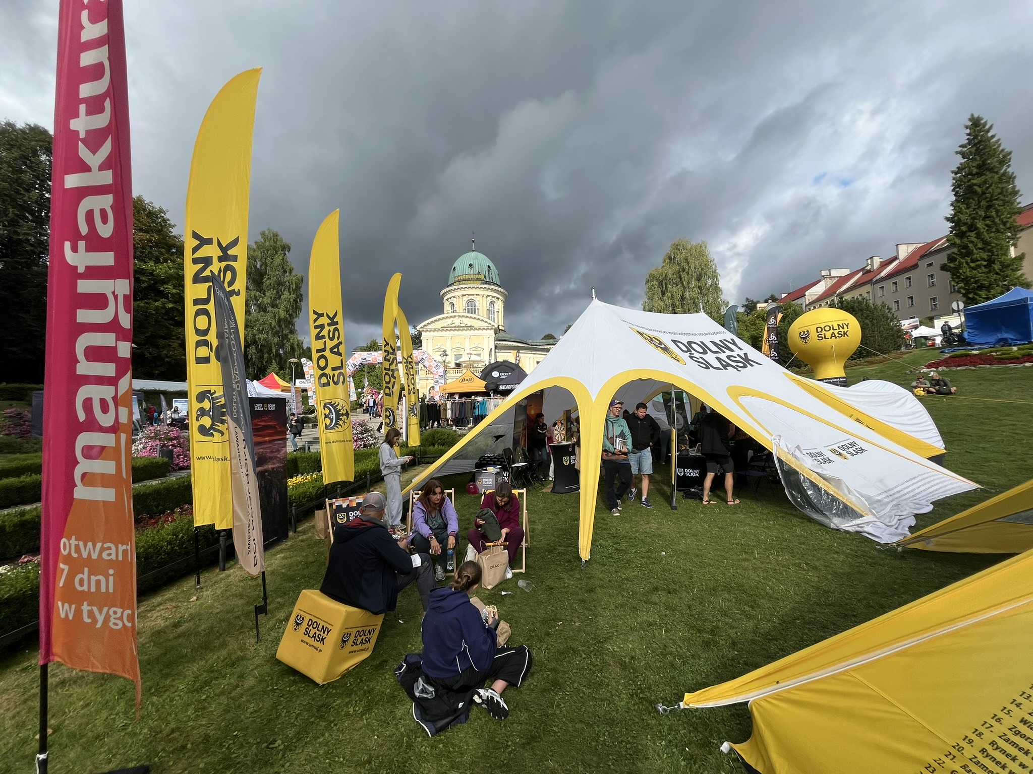 Widok na plenerową imprezę odbywającą się na trawniku pod zachmurzonym niebem. Na pierwszym planie widać dwie osoby siedzące na żółtych sześcianach i dwie siedzące na trawie. W tle stoją namioty, flagi i stoiska, a nad wszystkim góruje okazały budynek z kopułą Uzdrowiska Lądek Zdrój oraz drzewa. Na flagach, sześcianach i namiotach widnieje napis "DOLNY ŚLĄSK".