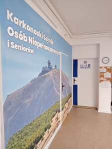 Fotografia przedstawia korytarz. Na lewej ścianie znajduje się duży plakat z widokiem na górę Śnieżkę, na której szczycie widać charakterystyczny budynek Obserwatorium Wysokogórskiego. Na plakacie widnieje napis w języku polskim: "Karkonoski Sejmik Osób Niepełnosprawnych i Seniorów". Po prawej stronie, na białych drzwiach, widać niebieski prostokąt z cyfrą "10" i tekstem "KLUB PACJENTA". Obok drzwi wisi tablica ogłoszeń.