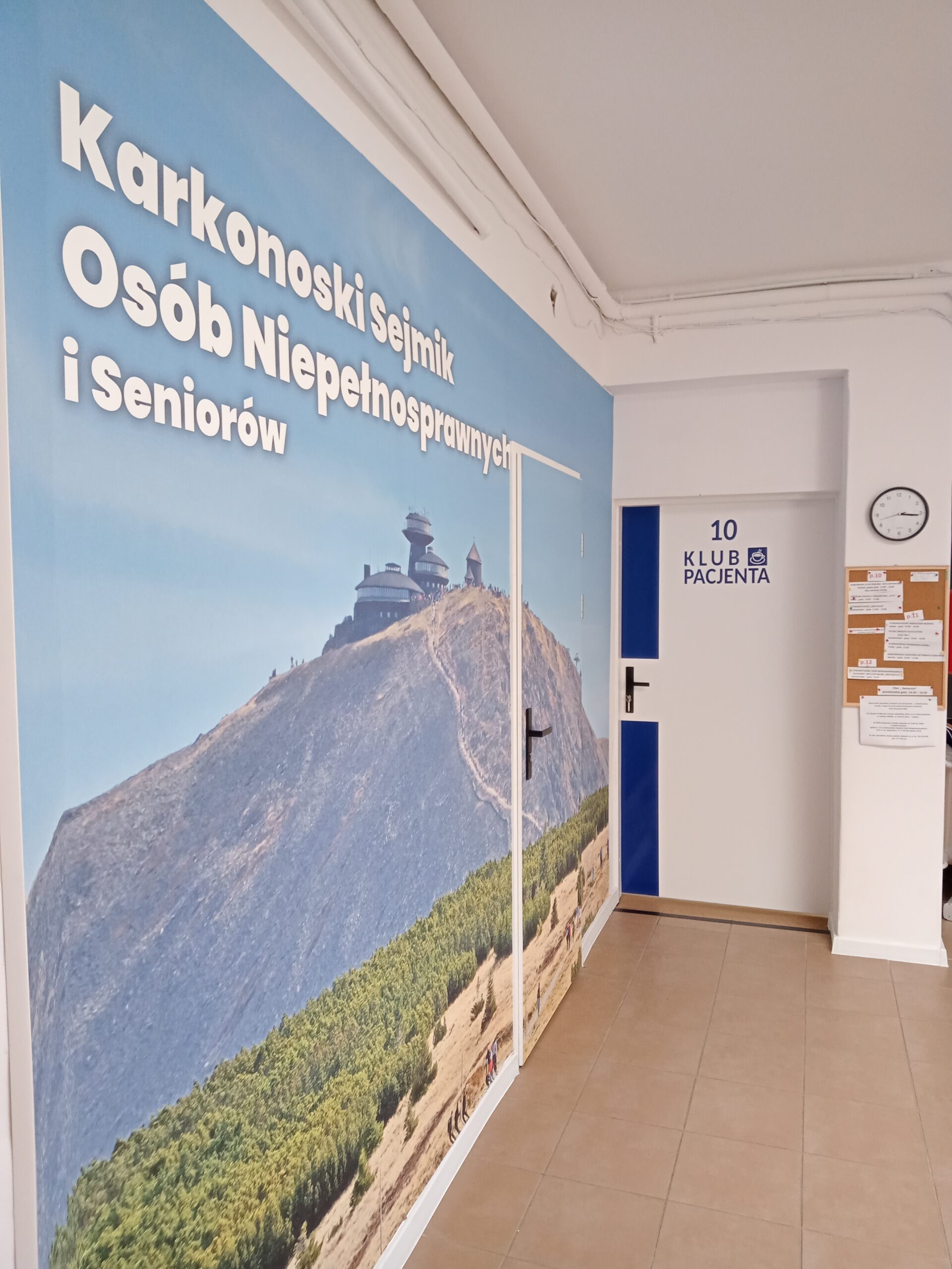 Fotografia przedstawia korytarz. Na lewej ścianie znajduje się duży plakat z widokiem na górę Śnieżkę, na której szczycie widać charakterystyczny budynek Obserwatorium Wysokogórskiego. Na plakacie widnieje napis w języku polskim: "Karkonoski Sejmik Osób Niepełnosprawnych i Seniorów". Po prawej stronie, na białych drzwiach, widać niebieski prostokąt z cyfrą "10" i tekstem "KLUB PACJENTA". Obok drzwi wisi tablica ogłoszeń.