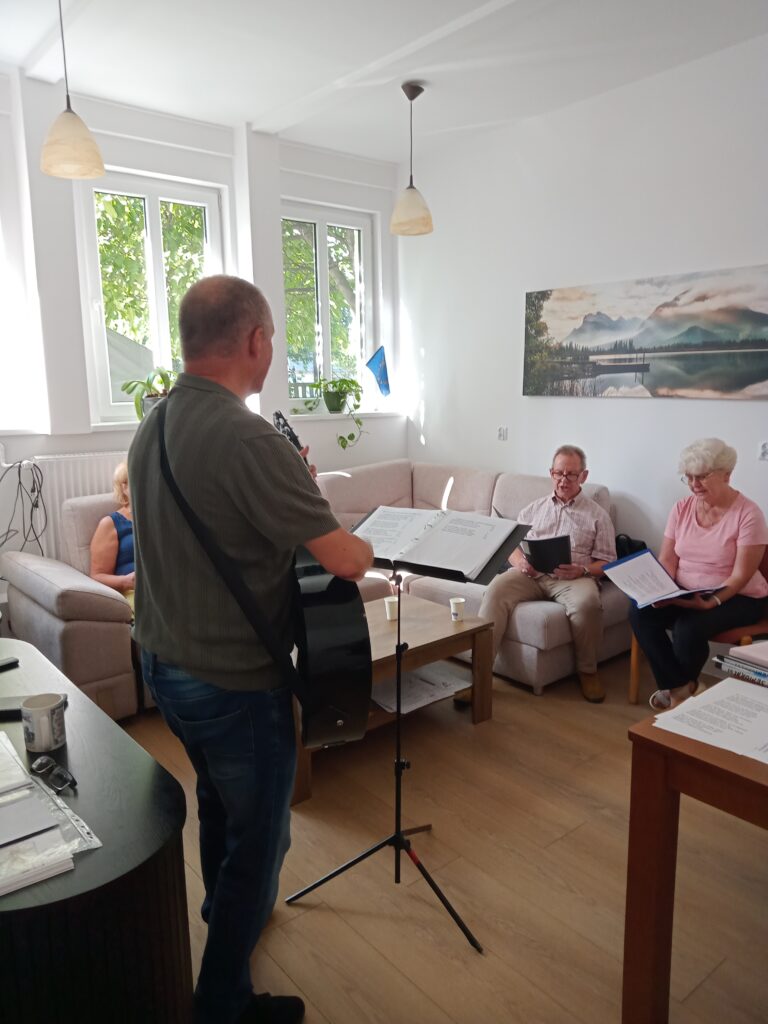 Fotografia przedstawia wnętrze pokoju. W centrum, tyłem do obiektywu, stoi mężczyzna grający na gitarze akustycznej, opierając się na pulpicie nutowym. W tle, na beżowej kanapie, siedzą trzy osoby — dwie kobiety i jeden mężczyzna. Wszyscy trzymają w rękach otwarte foldery z nutami lub tekstami piosenek, sugerując wspólne śpiewanie. W pokoju panuje jasne, naturalne oświetlenie wpadające przez duże okna. Na ścianie wisi obraz przedstawiający górski pejzaż.