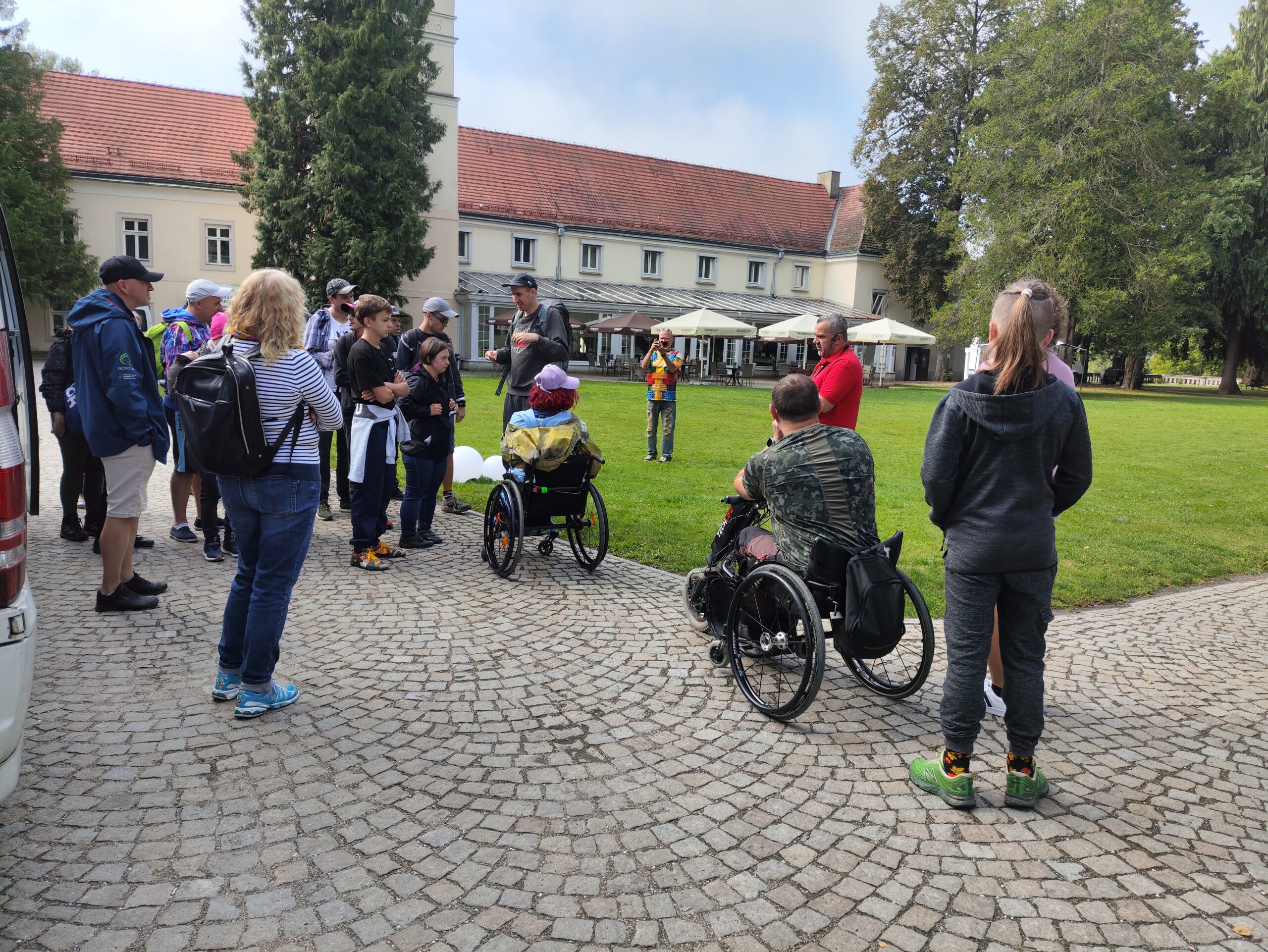 Zbiórka grupy osób na brukowanym podwórku przed dużym, jasnym zamkiem o spadzistym, czerwonym dachu. Z prawej strony widać dwóch dorosłych na wózkach inwalidzkich, a za nimi stoi dziewczyna w czarnej bluzie z kapturem. W tle, na trawniku, widać inne osoby.