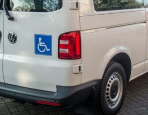 Obraz przedstawia zbliżenie na tył białego vana, z naklejką przedstawiającą symbol dostępu dla osób niepełnosprawnych, umieszczoną na lewym górnym rogu klapy bagażnika.