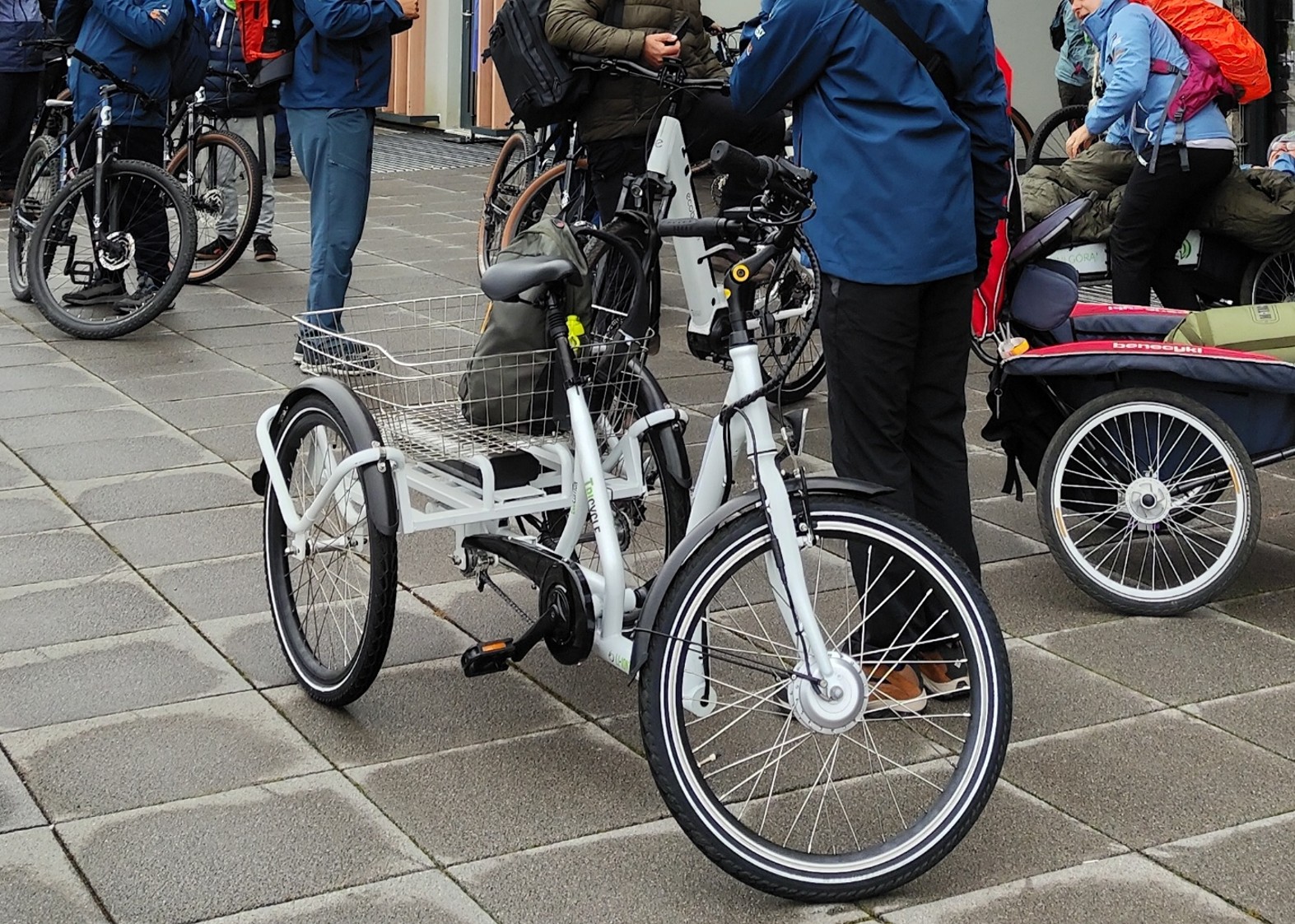 Rower trójkołowy z dużym koszem transportowym z tyłu stoi zaparkowany na chodniku wśród grupy osób ubranych w odzież turystyczną. W tle widoczne są inne rowery, w tym rowery górskie oraz przyczepka rowerowa. Osoby noszą plecaki i rozmawiają ze sobą, prawdopodobnie przygotowując się do wspólnej wyprawy rowerowej.