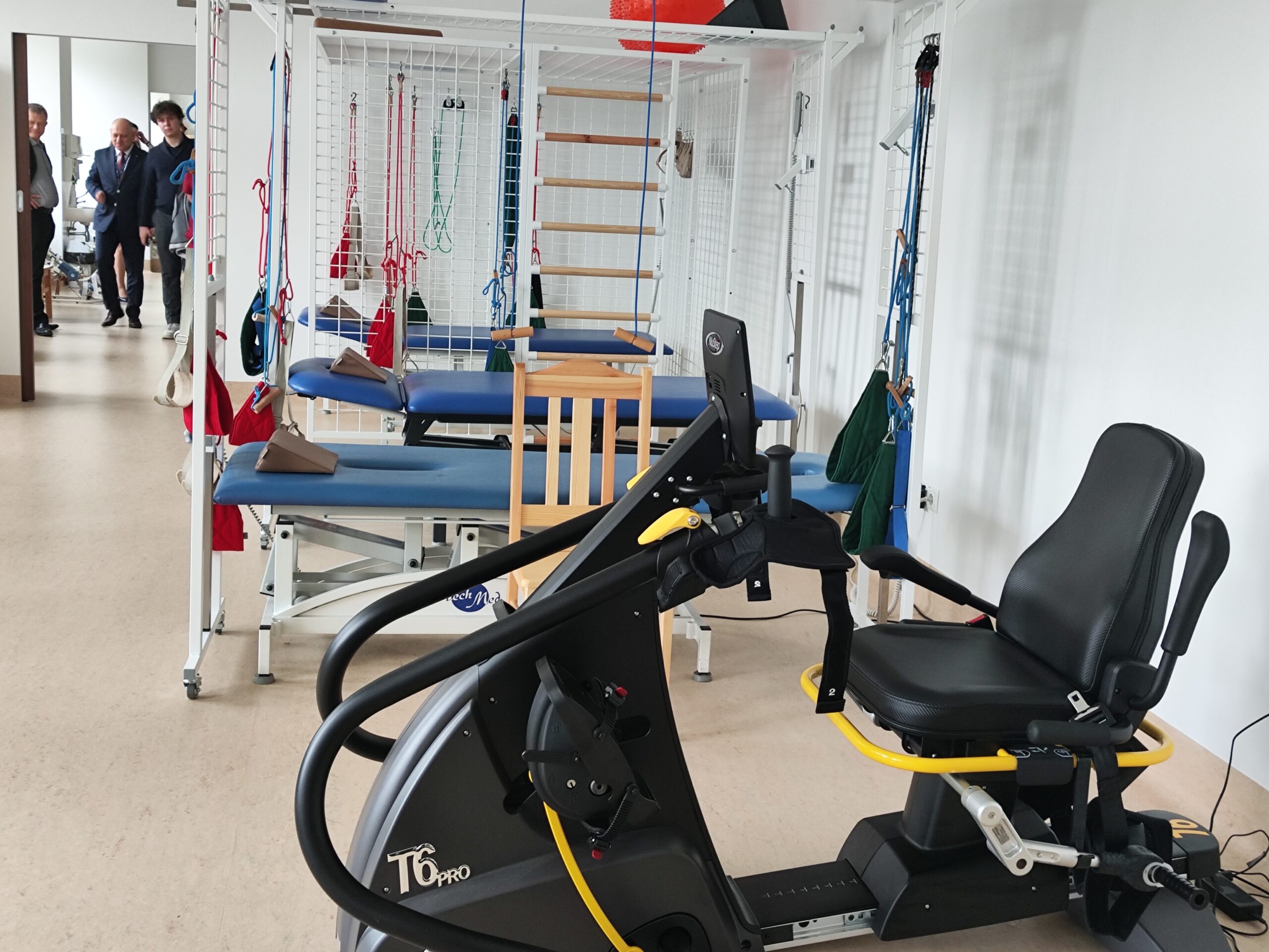 Zdjęcie przedstawia wnętrze sali do fizjoterapii lub rehabilitacji. Na pierwszym planie, z prawej strony, widoczny jest duży, czarny rower stacjonarny leżący (recumbent bike) z żółtymi akcentami, czarnym siedzeniem i uchwytami. W tle dominuje biały, metalowy stelaż, będący elementem wyposażenia do ćwiczeń. Do stelaża przymocowane są różnokolorowe taśmy, linki i uprzęże używane w kinezyterapii. Pomiędzy elementami stelaża znajdują się dwie niebieskie kozetki terapeutyczne, a na środku widoczne jest drewniane krzesło. Na jednej z półek stelaża leży czerwona piłka. Z lewej strony zdjęcia, w głębi, widać grupę osób w strojach wizytowych (mężczyźni w garniturach), stojących w korytarzu lub przejściu, patrzących w kierunku sali. Podłoga jest jasna. Oświetlenie jest jasne i równomierne.