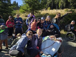 Grupa uśmiechniętych osób, w tym kilka siedzących lub stojących obok sprzętu do wędrówek z osobami z niepełnosprawnościami (wózek terenowy i plecaki), pozuje do wspólnego zdjęcia w słoneczny dzień na leśnej drodze. W tle widoczne są gęste drzewa i nasłoneczniona polana.