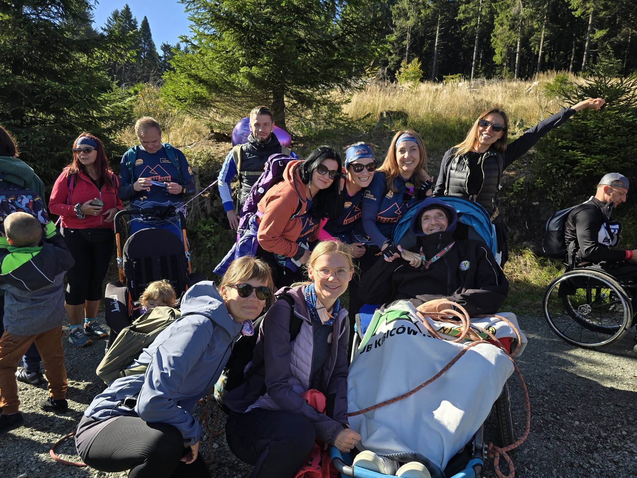 Grupa uśmiechniętych osób, w tym kilka siedzących lub stojących obok sprzętu do wędrówek z osobami z niepełnosprawnościami (wózek terenowy i plecaki), pozuje do wspólnego zdjęcia w słoneczny dzień na leśnej drodze. W tle widoczne są gęste drzewa i nasłoneczniona polana.
