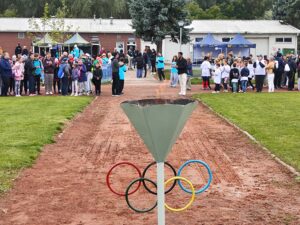 Obraz przedstawia pochodnię olimpijską w pierwszym planie, umieszczoną na środku ścieżki z czerwonej ziemi lub żwiru. Pochodnia ma stożkowaty kształt i znajduje się na białym słupie, u podstawy którego widoczne są sploty kół olimpijskich w kolorach czerwonym, czarnym, zielonym, żółtym i niebieskim. Nad pochodnią widać mały płomień. Ścieżka prowadzi w głąb obrazu, gdzie po obu stronach stoją grupy ludzi, obserwujących wydarzenie. Ubrani są w różne stroje, a część osób po prawej stronie ma na sobie białe koszulki z numerami lub symbolami, sugerujące udział w jakiejś konkurencji sportowej lub wydarzeniu. W tle widoczne są budynki o białych ścianach i szarych dachach, a przed nimi rozstawione są namioty i zadaszenia w kolorach niebieskim i zielonym. Pomiędzy budynkami i ludźmi rosną drzewa oraz widoczne są fragmenty trawiastych terenów. Całość sprawia wrażenie zawodów sportowych lub imprezy plenerowej.