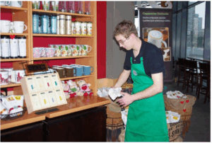 Mężczyzna w zielonym fartuchu, prawdopodobnie pracownik, jest zwrócony profilem i trzyma w dłoniach kawałek materiału, wykonując czynności porządkowe w obszarze z towarem na sprzedaż. W tle widać półki z kubkami, termosami i innymi produktami, a dalej za dużą szybą fragment wnętrza lokalu z siedzeniami i widok na zewnątrz. Na półce widoczne są również torebki herbaty i pluszowe misie.