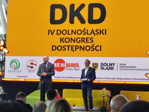 Dwóch mężczyzn stoi na scenie podczas IV Dolnośląskiego Kongresu Dostępności. W tle duży żółty ekran z napisem „DKD – IV Dolnośląski Kongres Dostępności” oraz logotypami organizatorów i partnerów wydarzenia, w tym Bonitum, Start Wrocław, Nie ma barier Dolny Śląsk i Urzędu Marszałkowskiego Województwa Dolnośląskiego. Przed sceną siedzi publiczność.