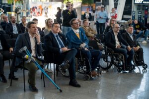 Grupa osób siedzi na krzesłach wewnątrz nowoczesnego budynku, podczas konferencji. W pierwszym rzędzie widocznych jest kilku mężczyzn, z których co najmniej trzech porusza się na wózkach lub używa kul. Uczestnicy są ubrani w stroje biznesowe lub półformalne i wydają się słuchać lub oglądać prezentację, która jest poza kadrem. W tle widać więcej osób stojących i siedzących, a także banery z logo i fragmenty informacji.