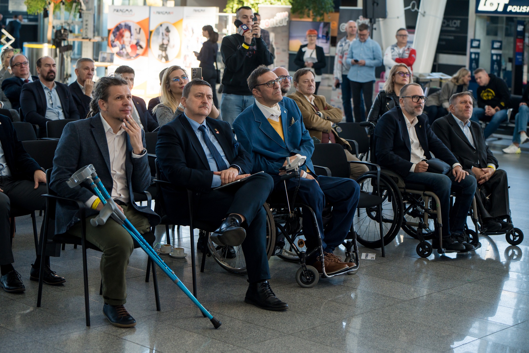 Grupa osób siedzi na krzesłach wewnątrz nowoczesnego budynku, podczas konferencji. W pierwszym rzędzie widocznych jest kilku mężczyzn, z których co najmniej trzech porusza się na wózkach lub używa kul. Uczestnicy są ubrani w stroje biznesowe lub półformalne i wydają się słuchać lub oglądać prezentację, która jest poza kadrem. W tle widać więcej osób stojących i siedzących, a także banery z logo i fragmenty informacji.