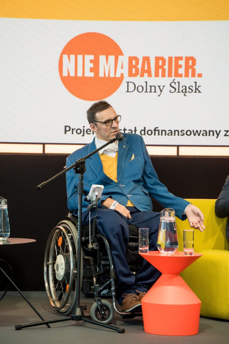 Grupa osób siedzi na krzesłach wewnątrz nowoczesnego budynku, prawdopodobnie podczas konferencji lub wydarzenia. W pierwszym rzędzie widocznych jest kilku mężczyzn, z których co najmniej trzech porusza się na wózkach inwalidzkich lub używa kul (widocznych przy krześle po lewej). Uczestnicy są ubrani w stroje biznesowe lub półformalne i wydają się słuchać lub oglądać prezentację, która jest poza kadrem. W tle widać więcej osób stojących i siedzących, a także banery z logo i fragmenty informacji.