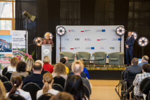 Wnętrze sali podczas oficjalnego wydarzenia. Na scenie stoi kobieta przemawiająca z mównicy, obok niej widoczne są reflektory oraz tło z logotypami instytucji rządowych, samorządowych i unijnych. Po prawej stronie sceny stoi elegancko ubrany mężczyzna. Przed sceną siedzi publiczność, w tym osoby na wózku. Po lewej stronie ustawiono roll-up z informacjami o Dolnośląskim Zespole Szkół w Biedrzychowicach.