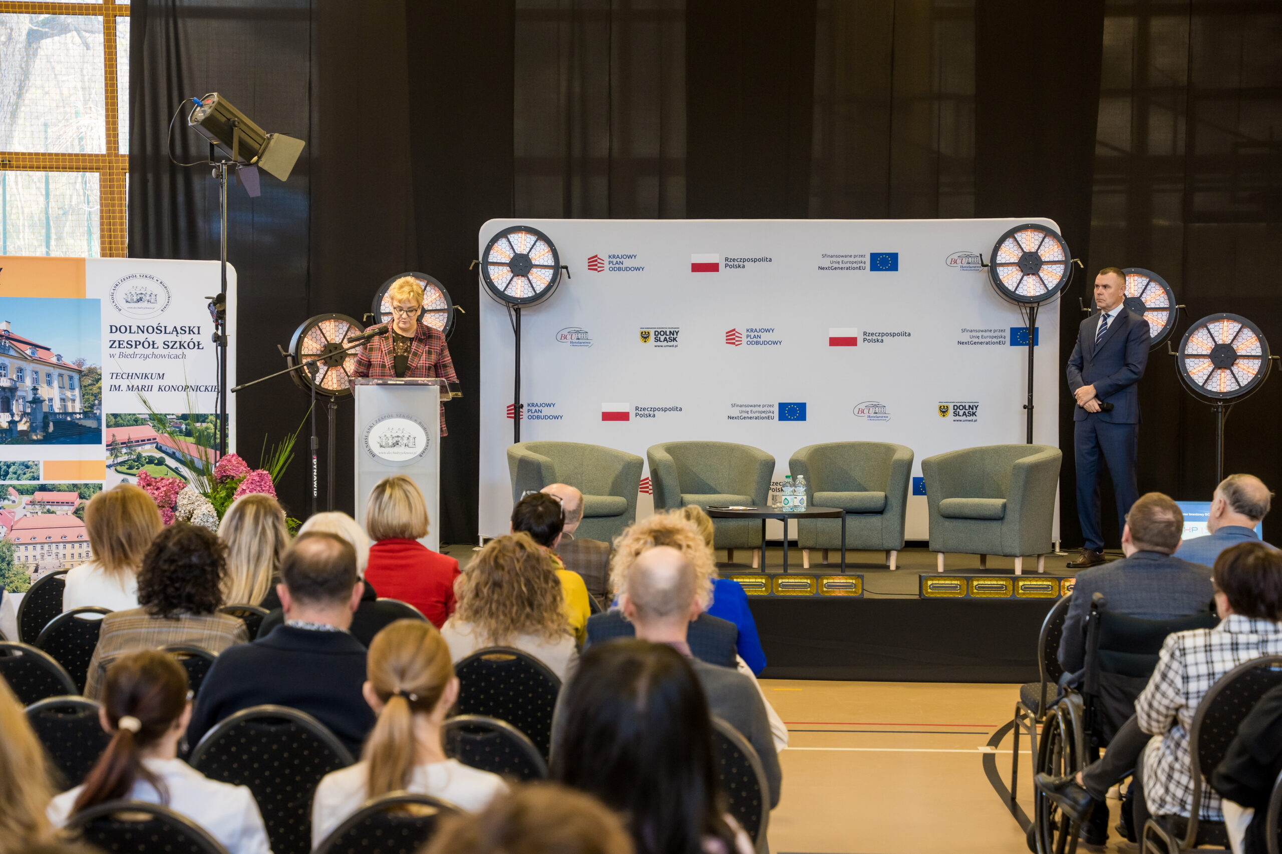 Wnętrze sali podczas oficjalnego wydarzenia. Na scenie stoi kobieta przemawiająca z mównicy, obok niej widoczne są reflektory oraz tło z logotypami instytucji rządowych, samorządowych i unijnych. Po prawej stronie sceny stoi elegancko ubrany mężczyzna. Przed sceną siedzi publiczność, w tym osoby na wózku. Po lewej stronie ustawiono roll-up z informacjami o Dolnośląskim Zespole Szkół w Biedrzychowicach.