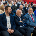 Grupa osób siedzących na widowni podczas oficjalnego wydarzenia. Na pierwszym planie trzech starszych mężczyzn w garniturach siedzi w pierwszym rzędzie, a za nimi widać wiele innych uczestniczek i uczestników słuchających wystąpień