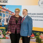 Kobieta w czerwono-czarnej kraciastej marynarce i mężczyzna w niebieskim, kraciastym garniturze z czerwonym krawatem pozują do zdjęcia. Stoją przed dużym banerem reklamowym Dolnośląskiego Zespołu Szkół im. Marii Konopnickiej w Biedrzychowicach, na którym widnieją zdjęcia zabytkowego budynku szkoły.