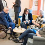 Grupa osób, w tym mężczyzna na wózku inwalidzkim oraz troje innych uczestników, prowadzi swobodną rozmowę przy kawie, siedząc w kąciku wypoczynkowym.