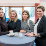 Cztery uśmiechnięte kobiety pozują do zdjęcia, stojąc przy małym stoliku z filiżankami kawy. W tle widoczne jest okno oraz na ścianie piktogramy ostrzegawcze dotyczące substancji niebezpiecznych.