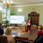 Kobieta w bordowym garniturze prezentuje slajd z mapą regionu podczas spotkania w eleganckiej sali konferencyjnej. Publiczność, składająca się z kilku osób, siedzi przy drewnianych stołach i słucha prezentacji.