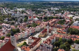 Zdjęcie z lotu ptaka przedstawiające rynek w Bolesławcu. W centrum widać historyczny ratusz z wysoką wieżą, otoczony kolorowymi, odrestaurowanymi kamienicami o czerwonych dachach. Po lewej stronie dominuje wieża kościoła pw. Wniebowzięcia NMP i św. Mikołaja. W tle rozpościera się widok na dalsze dzielnice miasta z dużą ilością zieleni i drzew pod jasnym niebem.
