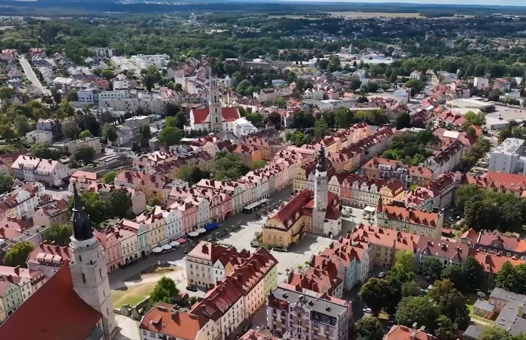 Zdjęcie z lotu ptaka przedstawiające rynek w Bolesławcu. W centrum widać historyczny ratusz z wysoką wieżą, otoczony kolorowymi, odrestaurowanymi kamienicami o czerwonych dachach. Po lewej stronie dominuje wieża kościoła pw. Wniebowzięcia NMP i św. Mikołaja. W tle rozpościera się widok na dalsze dzielnice miasta z dużą ilością zieleni i drzew pod jasnym niebem.