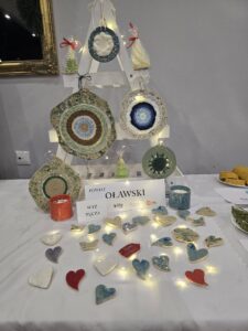 Zdjęcie przedstawia stoisko na festiwalu, prezentujące rękodzieło ceramiczne. Na białym obrusie leży duża liczba małych, glazurowanych serc oraz kilku innych kształtów (chmurki, ptaszki) w odcieniach bieli, czerwieni i niebieskiego, oświetlonych małą, ciepłą girlandą świetlną. Za nimi stoi biała, drewniana konstrukcja przypominająca drabinę lub choinkę, na której zawieszone są większe ceramiczne dekoracje w kształcie kół z wplecionym kolorowym sznurkiem (niebieski, zielony, brązowy) lub filcem, a także kilka małych, stożkowych świec świątecznych. Z boku widać również fragmenty innych produktów spożywczych (ciasta) oraz czerwoną i niebieską ceramiczną świecę/pojemnik.
