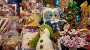 Zbliżenie na stoisko z rękodziełem świątecznym, głównie wykonanym na szydełku. W centralnej części widać wydzierganego na szydełku, uśmiechniętego bałwana w zielonej czapce i szaliku. Obok niego, nieco z tyłu, stoi większa, szydełkowa sowa w niebiesko-białym fraku. Po lewej stronie widać beżową, szydełkową choinkę. Po prawej, wśród dekoracji, znajduje się zielona, udekorowana szydełkowa choinka oraz ozdoby z suszonych materiałów i drobnych, czerwonych owoców.