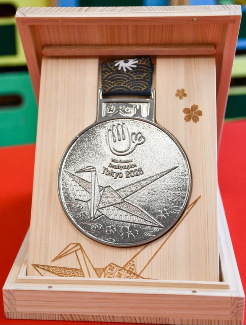 Na zdjęciu srebrny medal 25. Letnich Igrzysk Głuchych (Deaflympics) Tokyo 2025, umieszczony w jasnym, drewnianym etui. Na awersie medalu widnieje duży grawer papierowego żurawia (origami), logo Igrzysk Głuchych (symbol dłoni) oraz napis „Tokyo 2025”. Drewniana obudowa jest ozdobiona delikatnymi grawerami kwiatów wiśni oraz motywem żurawia w dolnej części.