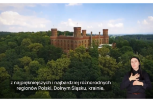 Pałac Marianny Orańskiej na wzgórzu w otoczeniu gęstych, zielonych drzew, sfotografowany z lotu ptaka. Na pierwszym planie, w prawym dolnym rogu, widoczna jest kobieta w ciemnym ubraniu, wykonująca gest języka migowego. W dolnej części ekranu znajduje się polski napis w białej ramce: "jeden z najpiękniejszych i najbardziej różnorodnych regionów Polski, Dolny Śląsk".