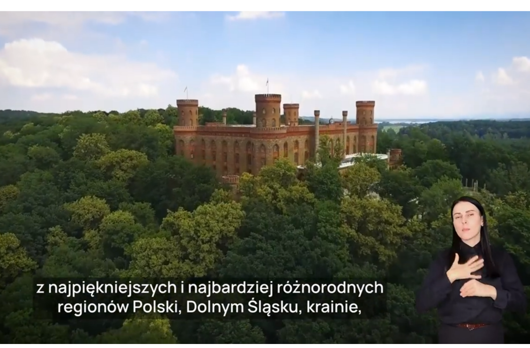 Pałac Marianny Orańskiej na wzgórzu w otoczeniu gęstych, zielonych drzew, sfotografowany z lotu ptaka. Na pierwszym planie, w prawym dolnym rogu, widoczna jest kobieta w ciemnym ubraniu, wykonująca gest języka migowego. W dolnej części ekranu znajduje się polski napis w białej ramce: "jeden z najpiękniejszych i najbardziej różnorodnych regionów Polski, Dolny Śląsk".