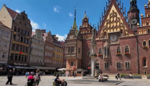 Słoneczny dzień na Rynku we Wrocławiu. Na pierwszym planie widać fragment brukowanego placu z przechodniami, w tym osobami poruszającymi się na wózkach inwalidzkich. W tle dominuje zabytkowy Stary Ratusz z charakterystyczną ceglaną fasadą, bogato zdobionym szczytem i zegarem astronomicznym. Obok Ratusza wznosi się kamienna Pręgierz. Po lewej stronie widoczne są kolorowe, zabytkowe kamienice, w tym jedna z szyldem restauracji Hard Rock Cafe. Niebo jest bezchmurne i błękitne.