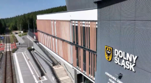 Widok z góry na elewację Dolnośląskiego Centrum Sportu w Jakuszycach. Na szarym fragmencie ściany widnieje herb Dolnego Śląska i napis „DOLNY ŚLĄSK”. Obok budynku znajduje się peron kolejowy, tory oraz gęsty las iglasty w tle