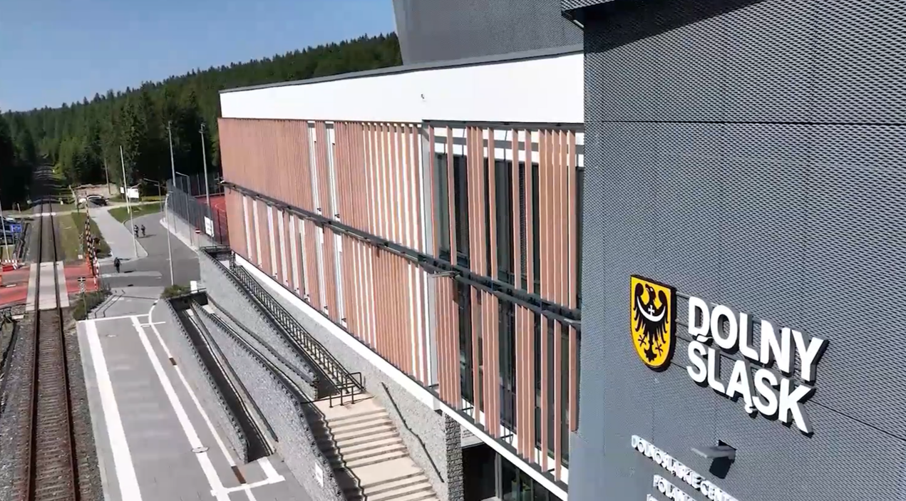 Widok z góry na elewację Dolnośląskiego Centrum Sportu w Jakuszycach. Na szarym fragmencie ściany widnieje herb Dolnego Śląska i napis „DOLNY ŚLĄSK”. Obok budynku znajduje się peron kolejowy, tory oraz gęsty las iglasty w tle