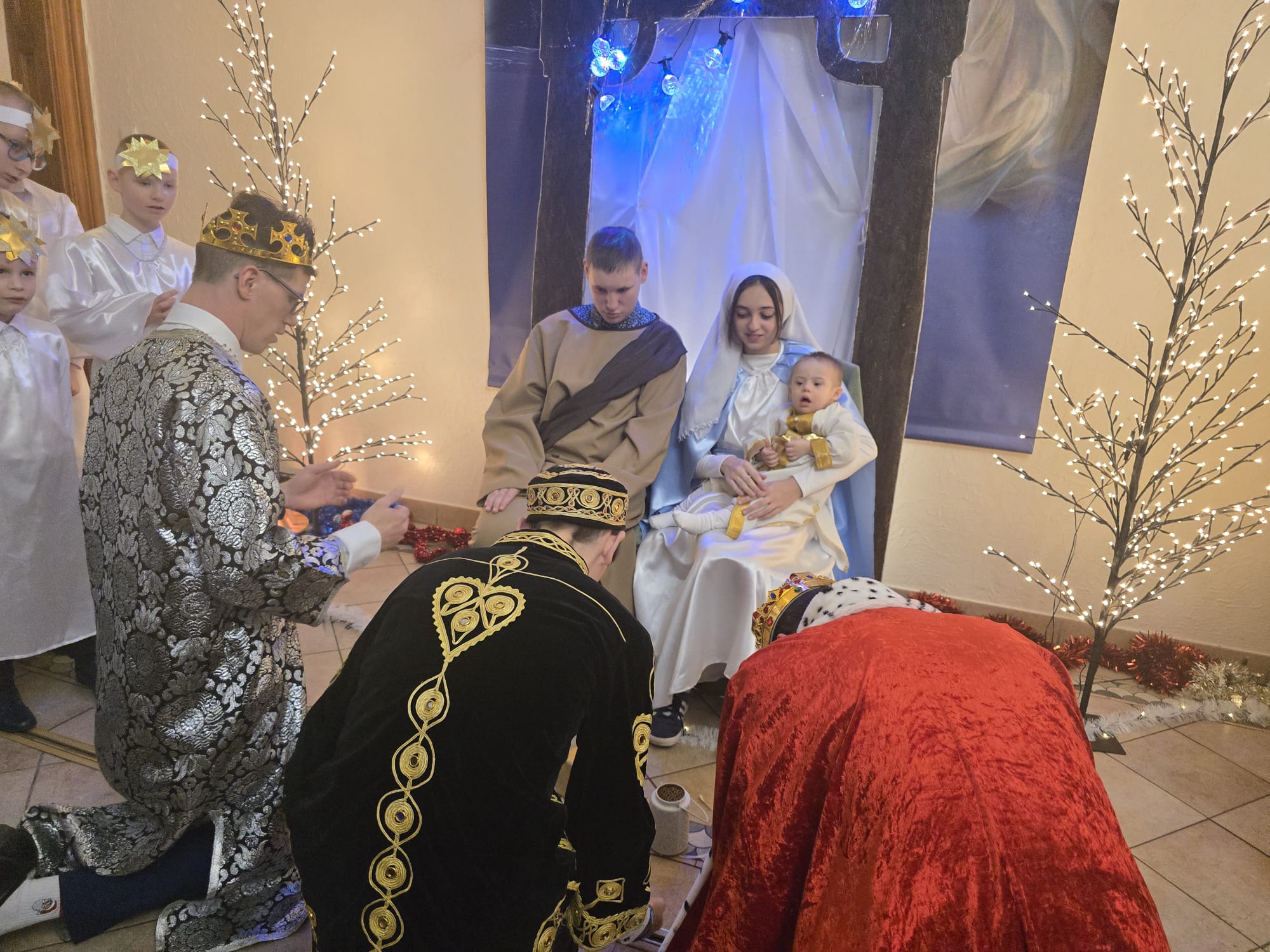 Grupa osób w kostiumach przedstawiająca scenę bożonarodzeniowych jasełek. W centralnym punkcie siedzi kobieta w biało-niebieskiej szacie, trzymająca na kolanach małe dziecko ubrane na biało. Obok nich siedzi młody mężczyzna w brązowej tunice. Przed nimi klęczą trzej mężczyźni przebrani za królów (mędrców) w bogato zdobionych szatach – złotej, czarnej oraz czerwonej. Po lewej stronie widać grupę dzieci w białych strojach z gwiazdami na opaskach, a całość dopełniają dekoracyjne, świecące drzewka w tle.