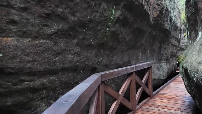 Zdjęcie przedstawia przejście turystyczne w labiryncie skalnym (charakterystyczne dla miejsc takich jak Błędne Skały czy Szczeliniec Wielki). Drewniany pomost prowadzi przez wąski korytarz wydrążony w piaskowcu, tworząc unikalny, niemal klaustrofobiczny klimat.